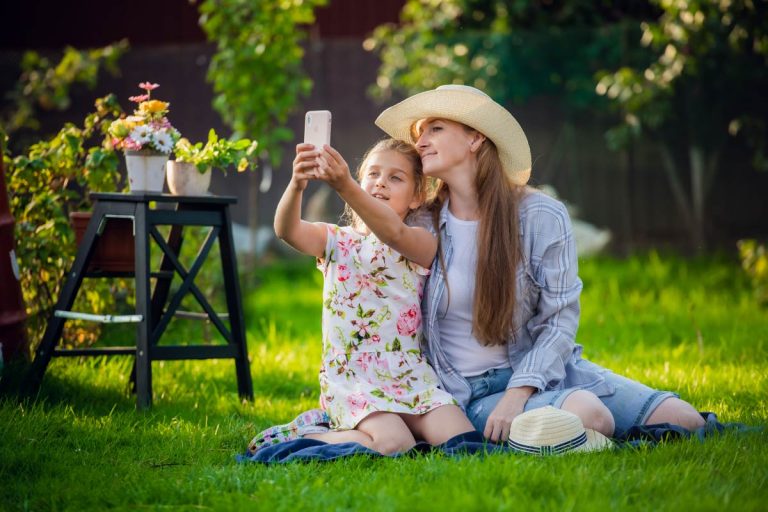 Smiling girl taking selfie with mother sitting on grass at backyard