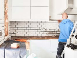 A man working in a new kitchen, a tiler applying tiles to the wall behind the cooker.